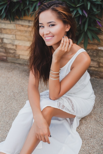 Giovane donna asiatica elegante che si siede sul pavimento in abito bianco boho, stile vintage, naturale, sorridente, felice, magro, vacanza tropicale, hotel