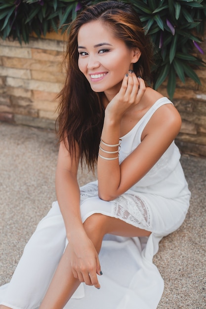 Young stylish asian woman sitting on floor in white boho dress, vintage style, natural, smiling, happy, skinny, tropical vacation, hotel