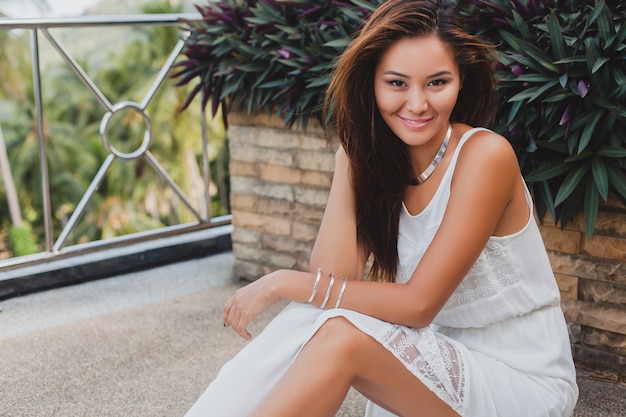 Young stylish asian woman sitting on floor in white boho dress, vintage style, natural, smiling, happy, skinny, tropical vacation, hotel