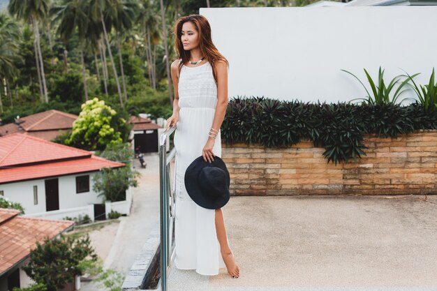 Young stylish asian woman in white boho dress, vintage style, natural, smiling, happy, tropical vacation, hotel, palm trees background