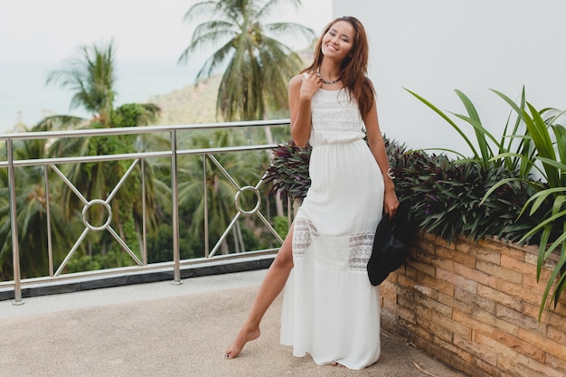 Young stylish asian woman in white boho dress, vintage style, natural, smiling, happy, tropical vacation, hotel, palm trees background