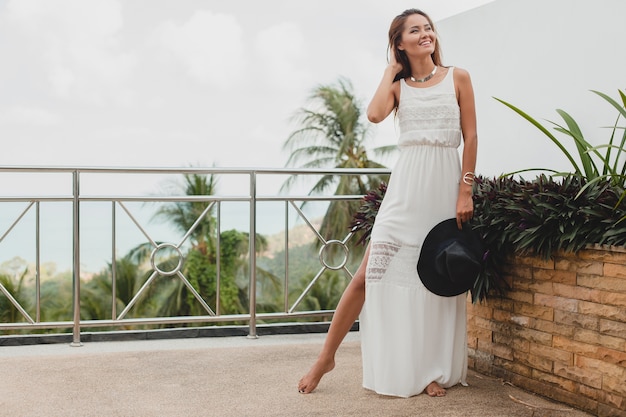 Free photo young stylish asian woman in white boho dress, vintage style, natural, smiling, happy, tropical vacation, hotel, palm trees background