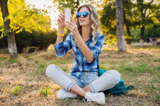 Молодая стильная привлекательная улыбающаяся белокурая женщина, сидящая в парке, летний повседневный стиль