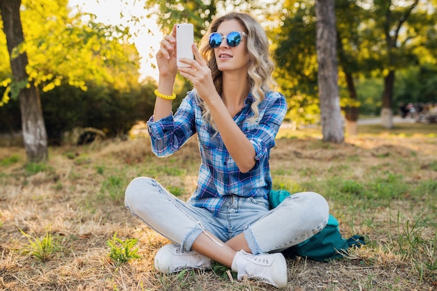 Молодая стильная привлекательная улыбающаяся белокурая женщина, сидящая в парке, летний повседневный стиль