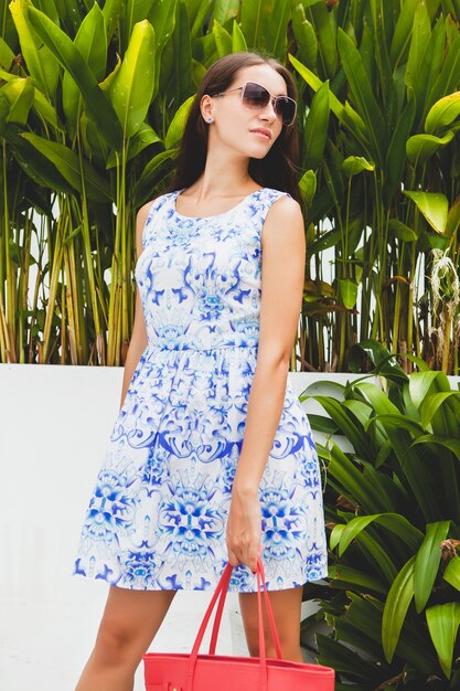 Young stylish beautiful woman in blue printed dress, red bag, sunglasses, happy mood, fashionable outfit, trendy apparel, smiling, summer, accessories