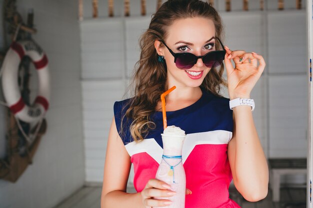 Young stylish beautiful woman in sea cafe, drinking cocktail smoothie, sunglasses, flirty, resort style, fashionable outfit, smiling, marine colors dress, anchor and lifebuoy on background, shocked
