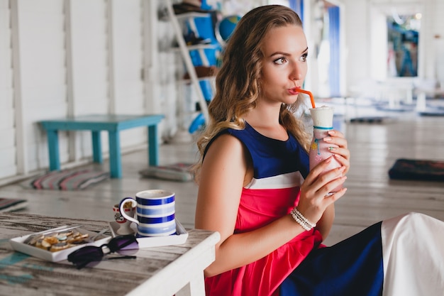 Young stylish beautiful woman in sea cafe, drinking cocktail smoothie, sunglasses, flirty, resort style, fashionable outfit, smiling, marine colors dress