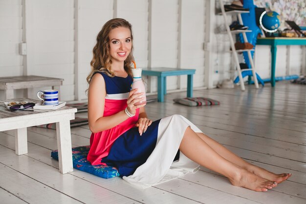 Young stylish beautiful woman in sea cafe, drinking cocktail smoothie, sunglasses, flirty, resort style, fashionable outfit, smiling, marine colors dress