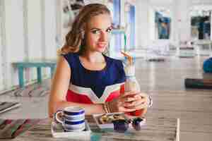 Free photo young stylish beautiful woman in sea cafe, drinking cocktail smoothie, sunglasses, flirty, resort style, fashionable outfit, smiling, marine colors dress