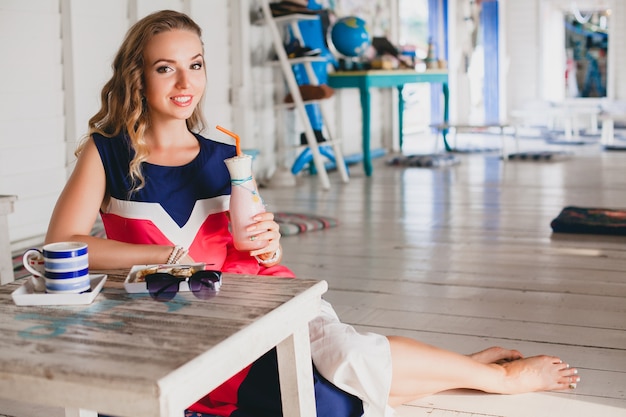 Young stylish beautiful woman in sea cafe, drinking cocktail smoothie, sunglasses, flirty, resort style, fashionable outfit, smiling, marine colors dress
