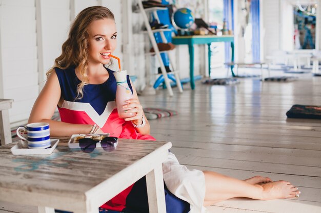 Young stylish beautiful woman in sea cafe, drinking cocktail smoothie, sunglasses, flirty, resort style, fashionable outfit, smiling, marine colors dress