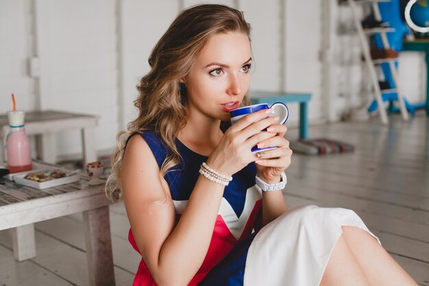 Young stylish beautiful woman in sea cafe, drinking hot cappuccino, resort style, fashionable outfit, smiling, marine colors dress, sitting on floor, vacation, relax