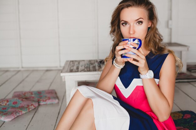 Young stylish beautiful woman in sea cafe, drinking hot cappuccino, resort style, fashionable outfit, smiling, marine colors dress, sitting on floor, vacation, relax