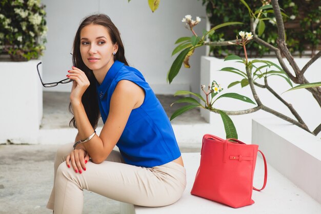 Young stylish beautiful woman, summer fashion trend, blue blouse, red bag, glasses, tropical villa resort, vacation, flirty