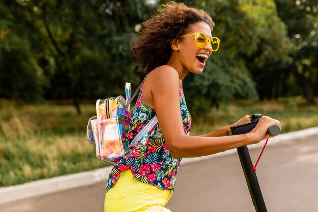 Young stylish black woman having fun in park riding on electric kick scooter in summer fashion style, colorful hipster outfit, wearing backpack and yellow sunglasses