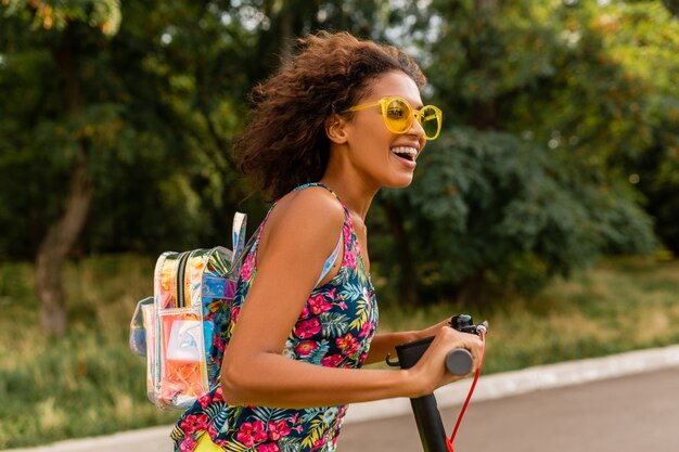 Young stylish black woman having fun in park riding on electric kick scooter in summer fashion style, colorful hipster outfit, wearing backpack and yellow sunglasses