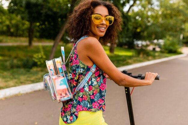 夏のファッションスタイル、カラフルなヒップスターの衣装、バックパックと黄色のサングラスを身に着けている電動キックスクーターに乗って公園で楽しんでいる若いスタイリッシュな黒人女性