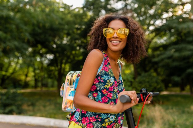 배낭과 노란색 선글라스를 착용, 여름 패션 스타일, 다채로운 힙 스터 복장에 전기 킥 스쿠터를 타고 공원에서 재미 젊은 세련된 흑인 여성