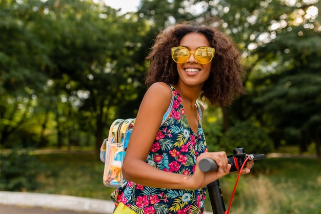 Young stylish black woman having fun in park riding on electric kick scooter in summer fashion style, colorful hipster outfit, wearing backpack and yellow sunglasses