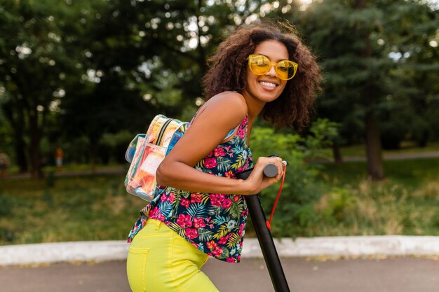 夏のファッションスタイル、カラフルなヒップスターの衣装、バックパックと黄色のサングラスを身に着けている電動キックスクーターに乗って公園で楽しんでいる若いスタイリッシュな黒人女性