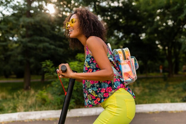 Young stylish black woman having fun in park riding on electric kick scooter in summer fashion style, colorful hipster outfit, wearing backpack and yellow sunglasses