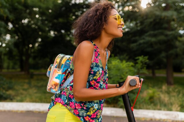 Giovane donna di colore alla moda che si diverte nel parco cavalcando scooter elettrico in stile moda estiva, vestito colorato hipster, indossando zaino e occhiali da sole gialli