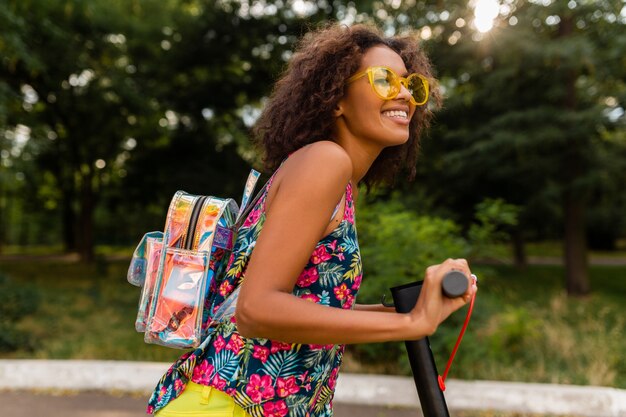 Молодая стильная темнокожая женщина веселится в парке, катается на электрическом самокате в стиле летней моды, в красочном хипстерском наряде, носит рюкзак и желтые солнцезащитные очки