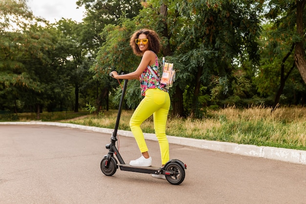 Young stylish black woman having fun in park riding on electric kick scooter in summer fashion style, colorful hipster outfit, wearing backpack and yellow trousers and sunglasses