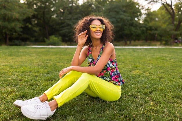 Young stylish black woman having fun in park summer fashion style, colorful hipster outfit, sitting on grass wearing yellow sunglasses and trousers, sneakers