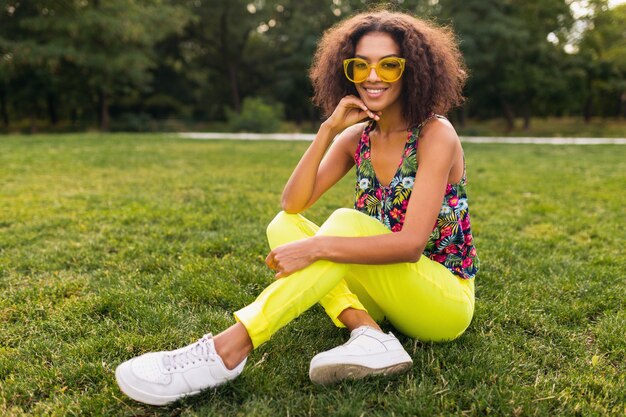 Young stylish black woman having fun in park summer fashion style, colorful hipster outfit, sitting on grass wearing yellow sunglasses and trousers, sneakers