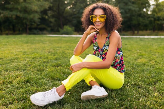 Молодая стильная темнокожая женщина веселится в парке летней моды, красочный хипстерский наряд, сидит на траве в желтых солнцезащитных очках и брюках, кроссовках