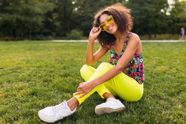 公園の夏のファッションスタイル、カラフルなヒップスターの衣装、黄色のサングラスとズボン、スニーカーを身に着けている草の上に座って楽しんでいる若いスタイリッシュな黒人女性