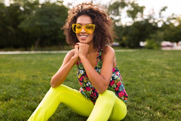 Молодая стильная темнокожая женщина веселится в парке в стиле летней моды, красочном хипстерском наряде, сидя на траве