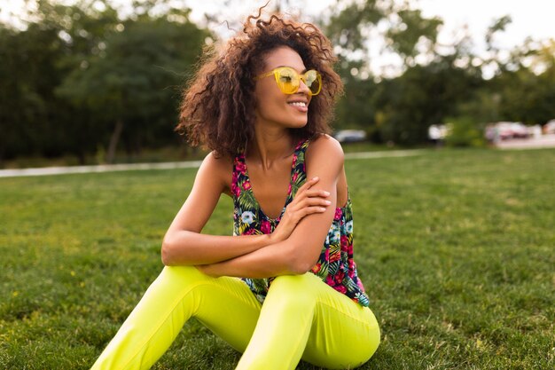 Young stylish black woman having fun in park summer fashion style, colorful hipster outfit, sitting on grass