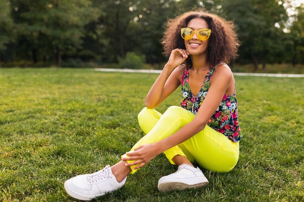 Молодая стильная темнокожая женщина веселится в парке в стиле летней моды, красочном хипстерском наряде, сидя на траве