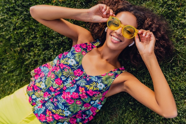 Free photo young stylish black woman listening to music on wireless earphones having fun lying on grass in park, summer fashion style, colorful hipster outfit, view from above