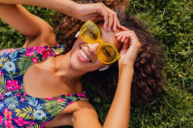 Young stylish black woman listening to music on wireless earphones having fun lying on grass in park, summer fashion style, colorful hipster outfit, view from above