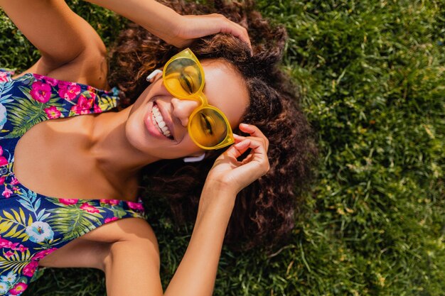 Young stylish black woman listening to music on wireless earphones having fun lying on grass in park, summer fashion style, colorful hipster outfit, view from above