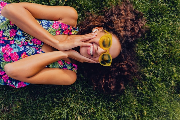 Giovane donna di colore alla moda che ascolta la musica sugli auricolari wireless divertendosi sdraiato sull'erba nel parco, stile di moda estiva, vestito colorato hipster, vista dall'alto