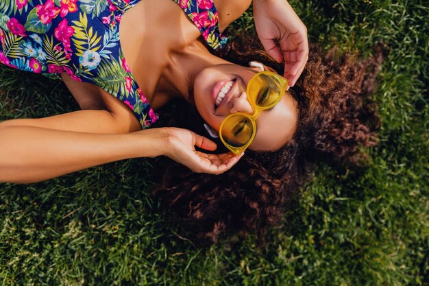 Young stylish black woman listening to music on wireless earphones having fun lying on grass in park, summer fashion style, colorful hipster outfit, view from above