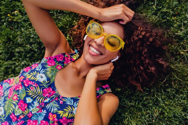 Giovane donna di colore alla moda che ascolta la musica sugli auricolari wireless divertendosi sdraiato sull'erba nel parco, stile di moda estiva, vestito colorato hipster, vista dall'alto