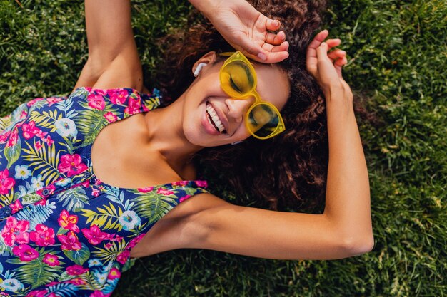 Young stylish black woman listening to music on wireless earphones having fun in park, summer fashion style, colorful hipster outfit, lying on grass, view from above