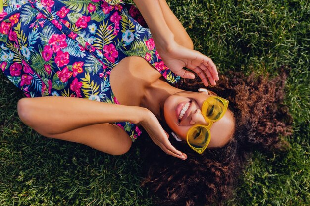 Young stylish black woman listening to music on wireless earphones having fun in park, summer fashion style, colorful hipster outfit, lying on grass, view from above