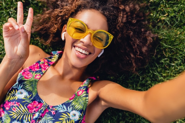 Молодая стильная темнокожая женщина, слушающая музыку на беспроводных наушниках, веселится в парке, в летнем модном стиле, красочный хипстерский наряд, лежа на траве, вид сверху