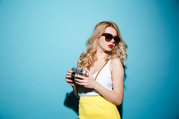 Giovane donna bionda alla moda in occhiali da sole che tengono retro macchina fotografica