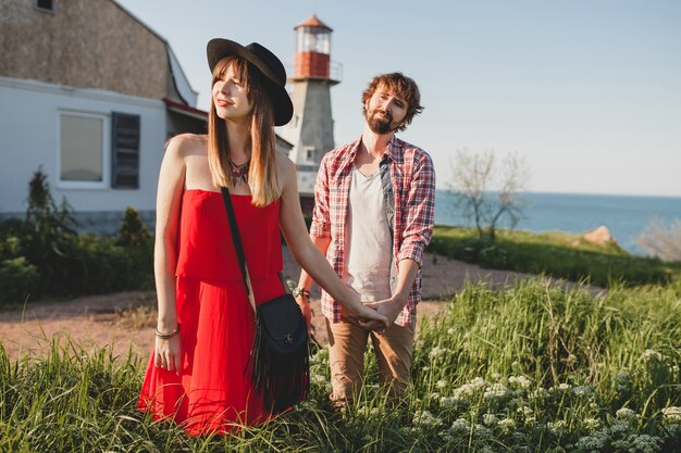 Young stylish couple in love in countryside, indie hipster bohemian style, weekend vacation, summer outfit, red dress, green grass, holding hands, smiling