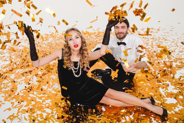 Young stylish couple in love, sitting on floor, throwing golden confetti