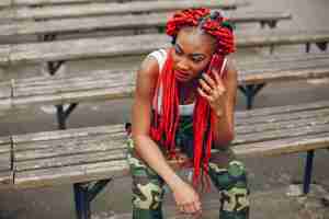 Free photo a young and stylish dark-skinned girl with red dreads sitting in the summer park