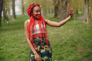 Free photo a young and stylish dark-skinned girl with red dreads walking in the summer park with phone