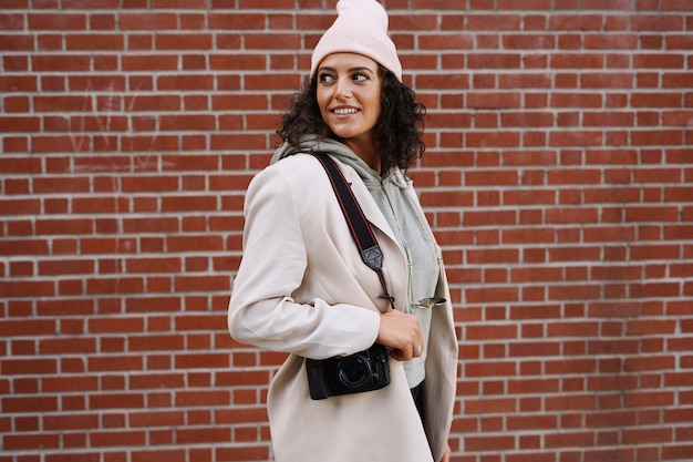 Young stylish female photographer posing against brick wall in the city